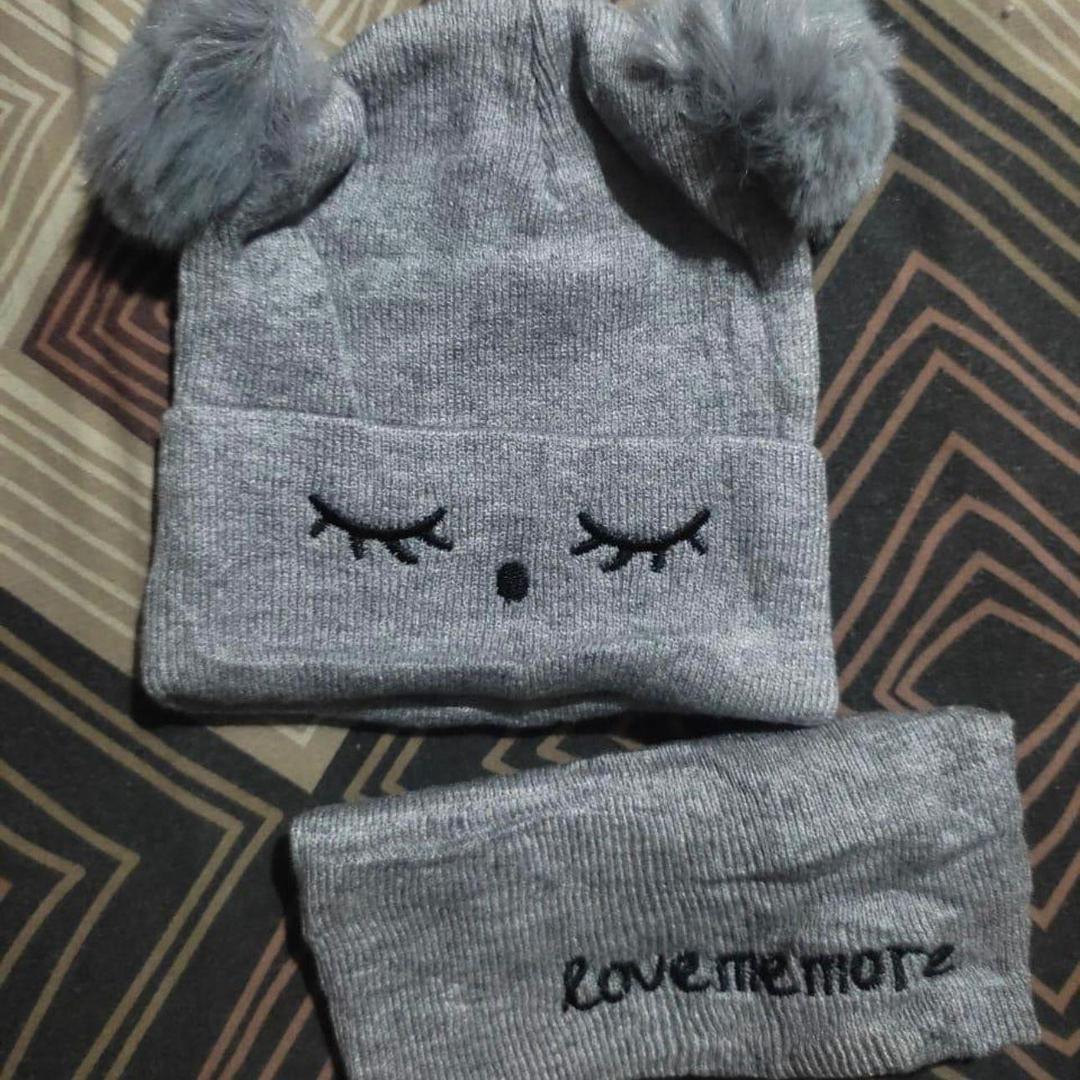 Gray knit baby hat with pom-pom ears and closed eyes, matching headband on patterned fabric.