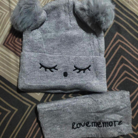 Gray knit baby hat with pom-pom ears and closed eyes, matching headband on patterned fabric.