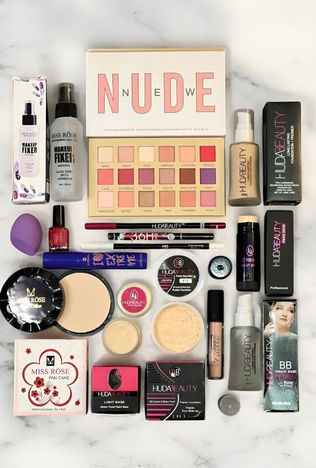 Flat lay of assorted makeup products including Huda Beauty eyeshadow palette, foundation, primer, Miss Rose makeup fixer spray, powders, brushes, and cosmetic tools on a marble background.