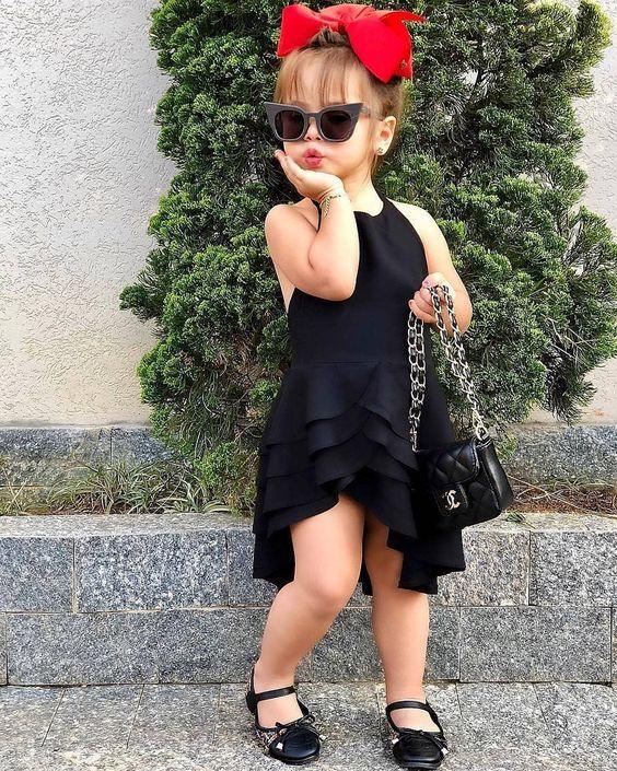 Stylish little girl in black dress, sunglasses, red bow, and purse posing outdoors