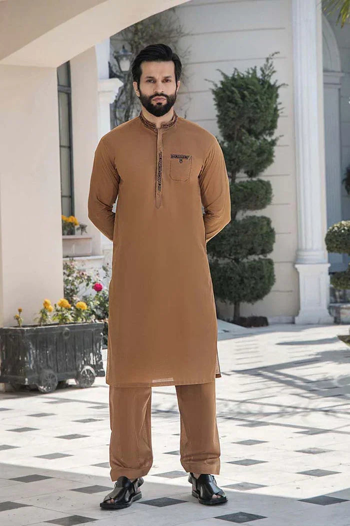 Man wearing brown shalwar kameez outfit, standing outdoors by flowering plants and topiary.